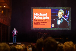 Jacob Yarrow standing on stage in spotlight in front of audience with "Michael Feinstein, Because of You" screen beside him